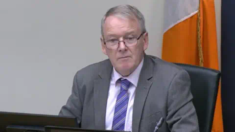 Oireachtas  Brian Stanley is sitting in a committee. He's wearing black glasses and a grey suit with a white shirt and a purple tie. The Irish tricolour flag is behind him.